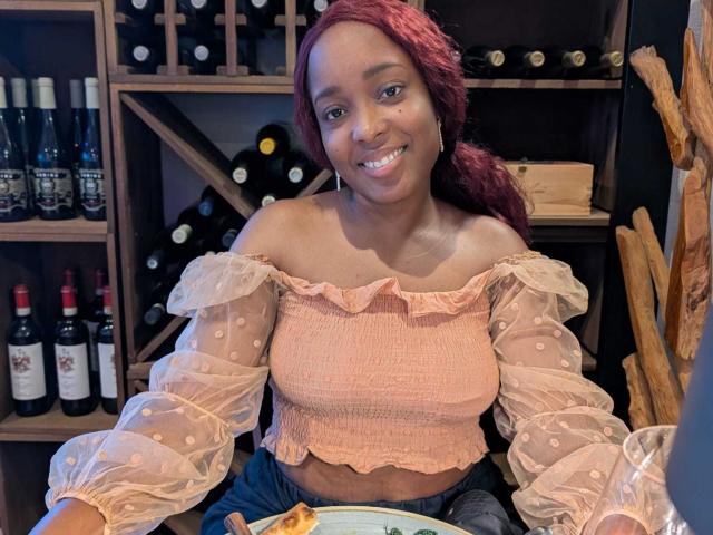ChelseyeBonny with dark skin, red hair in a ponytail, wearing a pink off-shoulder polka dot top, smiling, sitting at a table with a plate of food.