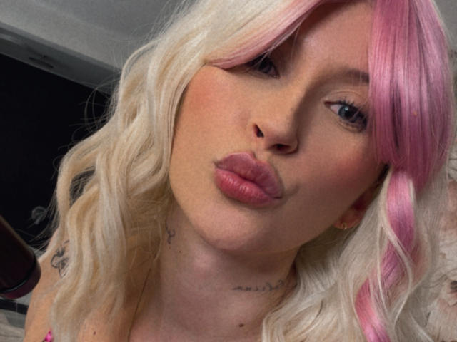 Emmyy with pink and blonde wavy hair, pouty pink lips, tattoos on neck, and blue eyes, making a kissing face. Her tits are slightly visible under her top.