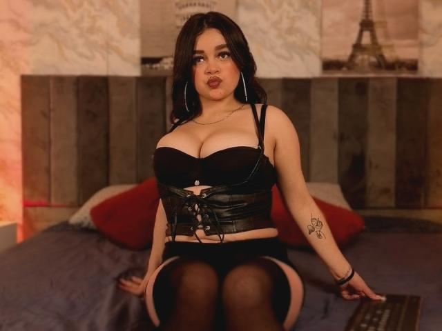 FabiValdez with large boobs, black leather corset, black garter, and stockings, sitting on a bed, looking up with pouty lips, wearing hoop earrings, tattoo on right arm.