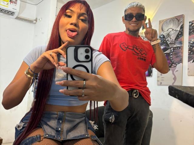 MegaSexyHot with long red hair, tongue out, blue crop top, denim skirt, taking selfie, showing ass with fishnet, standing next to MegaSexyHot in red tee, black pants.