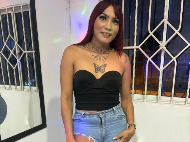 MegaSexyHot with red hair, black strapless top, butterfly tattoo on chest, denim shorts, tattoos on neck and arm, smiling, standing in front of white window with metal bars.