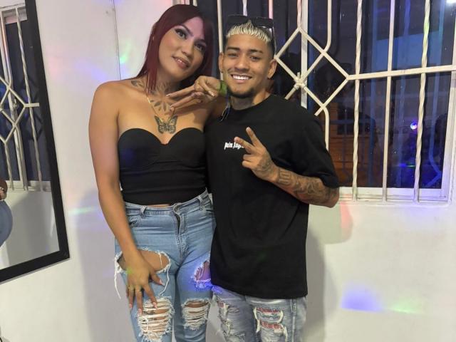 MegaSexyHot with red hair and tattoos, in a black strapless top and ripped jeans, stands next to MegaSexyHot with bleached hair, tattoos, and a black shirt, smiling.