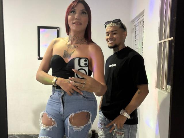 MegaSexyHot with red hair, black strapless top, and ripped jeans stands next to MegaSexyHot in black shirt and jeans, holding phone. Both smiling, tattoos visible.