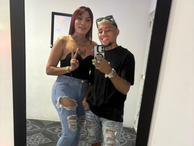MegaSexyHot with red hair, tattoos, black strapless top, and ripped jeans stands next to MegaSexyHot with blonde undercuts, black t-shirt, and ripped jeans, taking a mirror selfie.