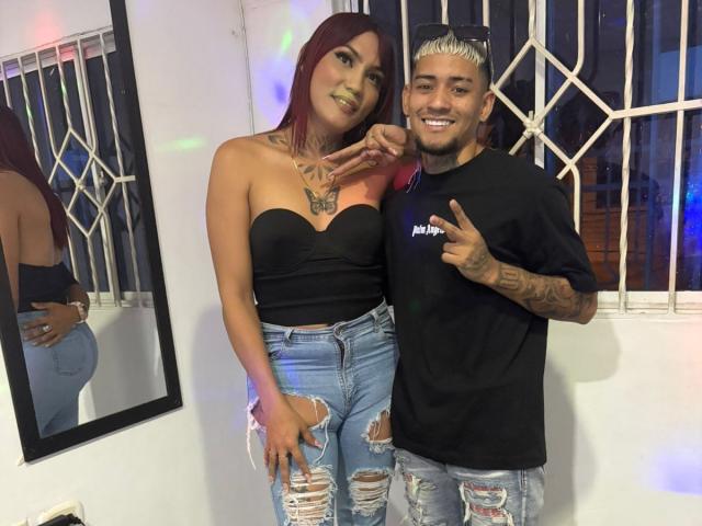 MegaSexyHot with medium tits and green lipstick, wearing a black strapless top and ripped jeans, stands next to MegaSexyHot with tattoos, black shirt, and bleached hair, flashing.