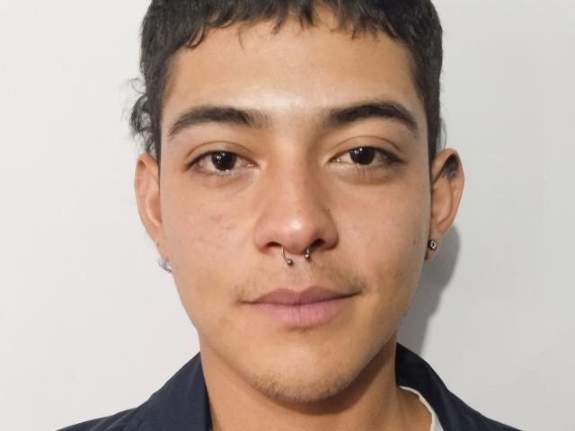 JackieThomas has a nose ring, small earrings, and a slight mustache, with short curly black hair, and a small smile, wearing a dark shirt.