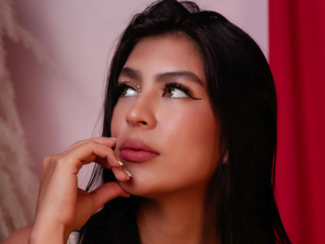SamanthaEva69 has long black hair, light brown skin, and is looking up with her finger near her mouth. Her eyelashes are long, and she has pink lipstick.