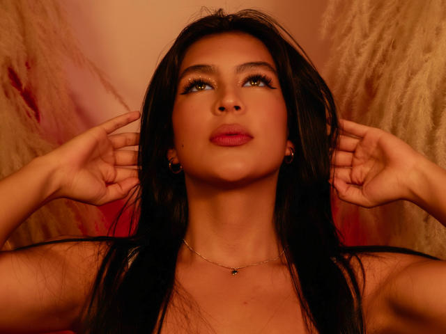SamanthaEva69 with long black hair, green eyes, and red lipstick, hands on ears, looking up, gold necklace, warm brown and red lighting, boho style, serious expression.