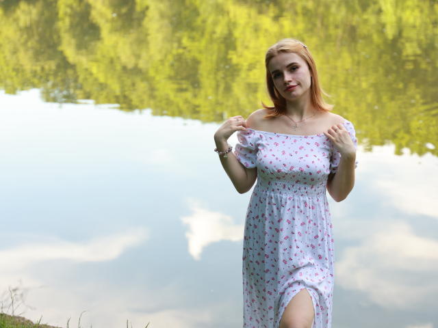 MargaritaKis with pink-haired bob, off-shoulder white dress with pink floral print, holding dress straps, standing by calm water, reflection shows green trees, subtle smile, thigh-high dress slit.