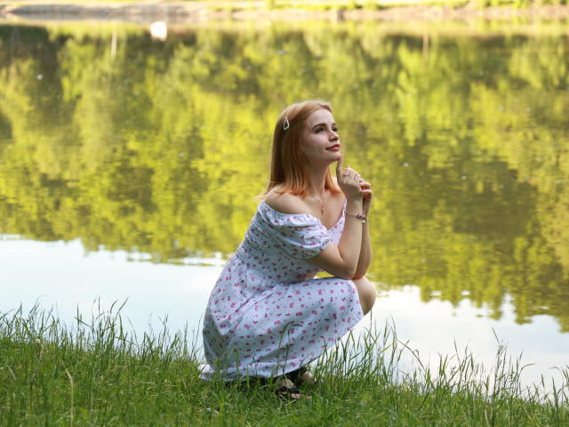 MargaritaKis, squatting on grass, wearing a white floral dress, off-shoulder, holding a straw, red hair, reflective green water, small gold hair clip, soft smile, summer.