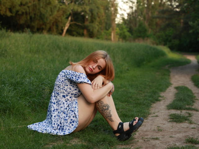 MargaritaKis, sitting on grass, wearing off-shoulder blue floral dress, with hand on chin, legs bent, showing thigh tattoo, looking at camera, in daylight, blonde hair, bracelet.