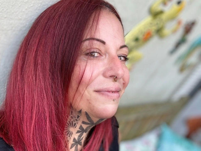 Miss-Ruby-Rubens-hot with red hair, nose ring, lip piercing, and tattooed neck, smiling at camera, showing her pierced ass in the background. Blurred yellow toy in the background.