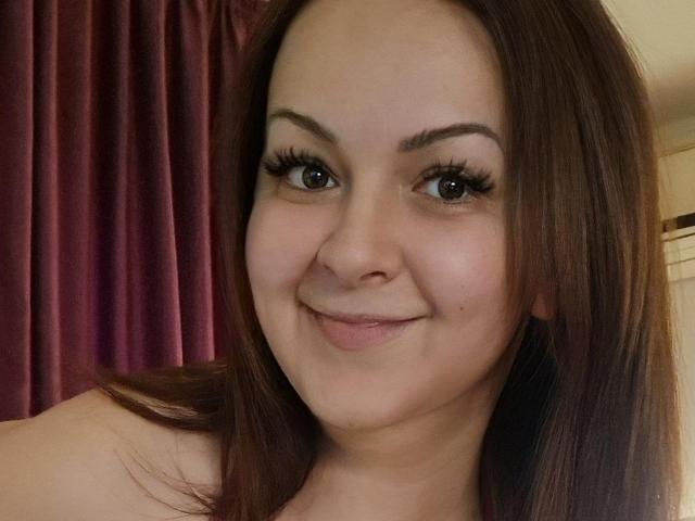 LoraHazel tiene el cabello castaño, cejas pobladas, pestañas largas y una ligera sonrisa que muestra sus pequeños senos y su piel clara. Sus ojos son marrones y está mirando a la cámara..