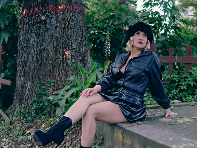GiuliaAddams with blonde hair, black fur hat, red lipstick, black leather dress, and black boots, sitting on a stone bench, legs crossed, showing her bare thighs and black panties.