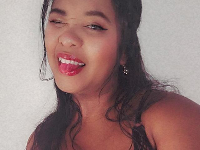 LilithAthenea's face, winking with her tongue out, showing off her red lipstick and white teeth, has wet black hair and small hoop earrings, against a plain white background.