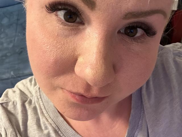 PussyDolly69's face, showing her brown eyes with long eyelashes, slightly parted pink lips, and a neutral expression, wearing a gray shirt with a V-neck, close-up selfie.