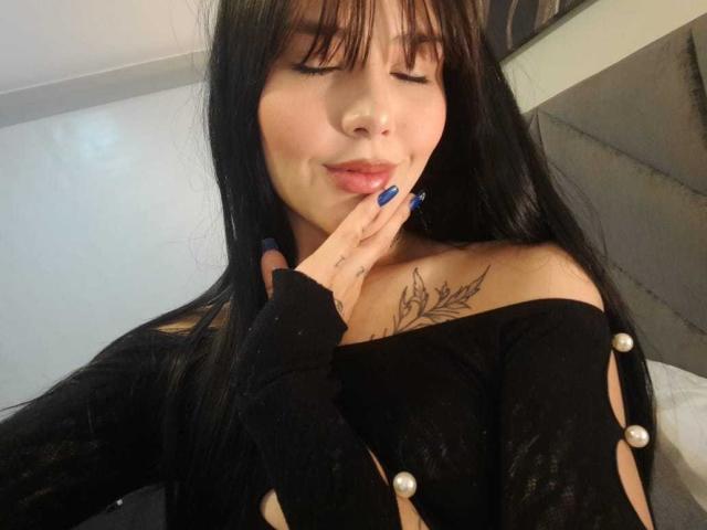 ChloeSantory with long black hair, closed eyes, and blue nails, touching her lips, wearing a black off-shoulder top with pearl buttons, showing a tattoo on her chest.
