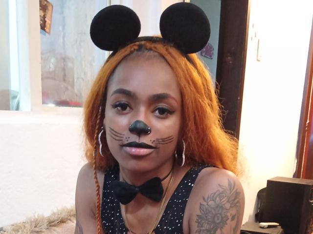 KatalinaCandy has orange hair, black mouse ears, black nose, whisker makeup, hoop earrings, black sparkly top, bowtie, tattooed arm, and is looking at the camera.