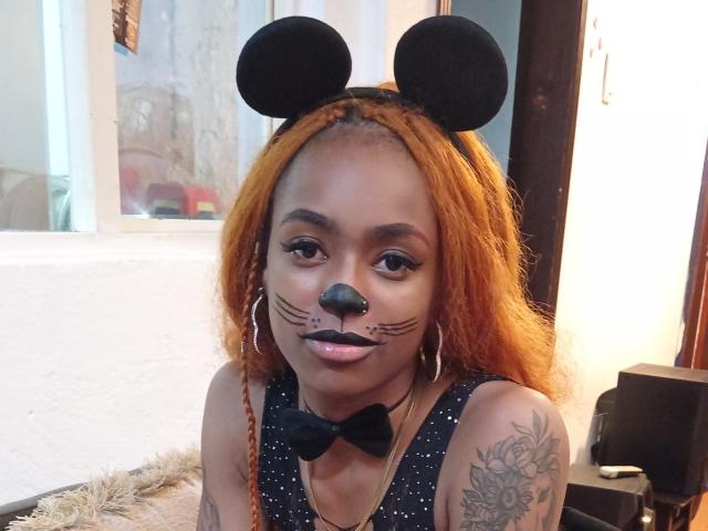KatalinaCandy with orange braids, black Minnie Mouse ears, black nose, whisker makeup, black bowtie, and hooped earrings, showing tattoos on her right arm.