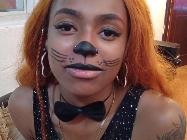 KatalinaCandy with orange braids, black nose, whisker tattoos, and black lipstick, wearing a black bowtie and silver hoop earrings, smiling at the camera.