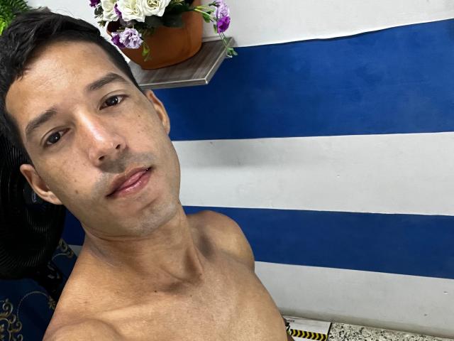 ManoloCardona, shirtless, with short black hair and slight stubble, brown eyes, and a relaxed expression, in front of a blue and white striped background with a flower pot.