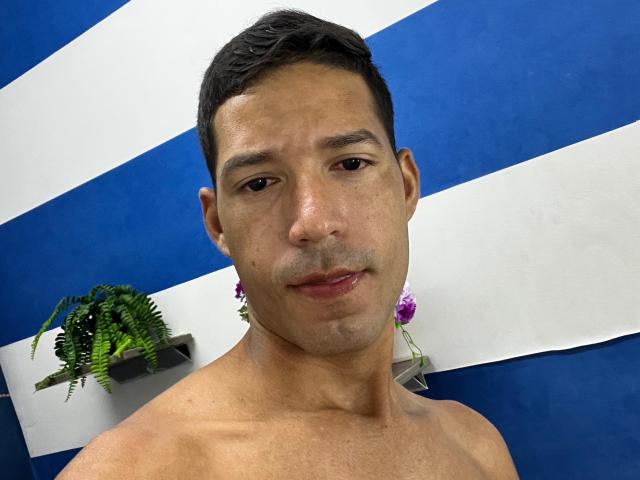 Manolo Cardona sans chemise, avec des cheveux noirs courts et une légère barbe de trois jours, regardant la caméra avec un léger sourire, mur rayé bleu et blanc en arrière-plan, petite plante de fougère sur la gauche..
