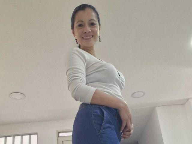 Melissa Baud mit dunklem Haar zu einem Dutt gebunden, trägt ein weißes Oberteil mit langen Ärmeln und blaue Jeans, lächelt, hat eine Hand auf dem Bauch, trägt Ohrringe und steht in einem hellen Raum..