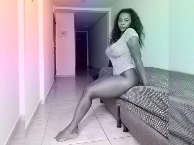 MacarenaBoobs with curly hair, wearing a tight gray shirt, sitting on a gray couch, showing off her big ass and long legs, in a tiled hallway, barefoot.