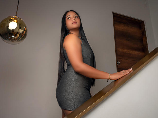 MiaLuz with long black hair, wearing a tight gray dress, standing on a staircase, looking back, showing her ass, in a room with a wooden door and a hanging light.