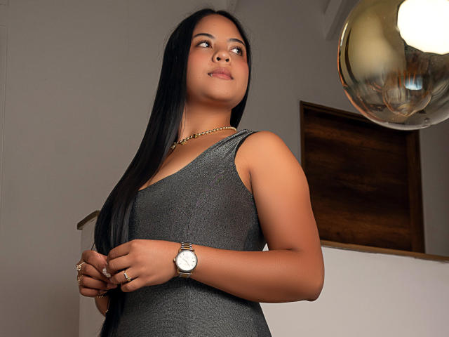 MiaLuz has long black hair, a tan skin tone, and is wearing a gray sleeveless dress with a gold chain necklace and a silver watch. She looks up with a slight smile.