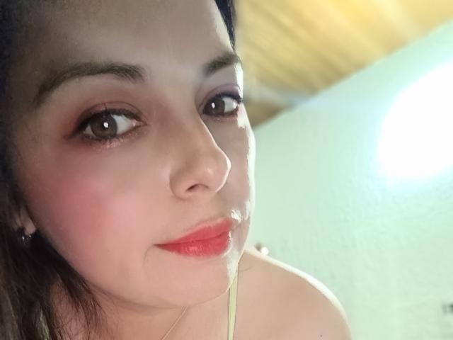Seher's face, showing a slight smile with red lips, brown eyes, and dark eyebrows, wearing a necklace, with a subtle glow on her fair skin.