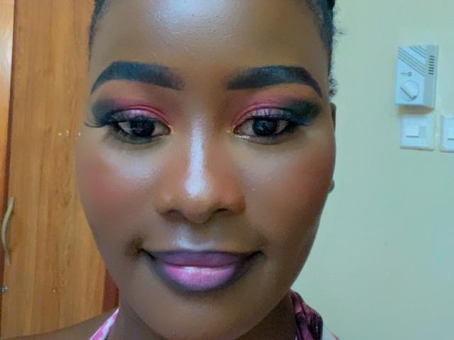 ElaraForYou has dark brown skin, pink and purple eyeshadow, thick black eyebrows, long black eyelashes, glossy pink lips, and a small smile, with her hair in a bun.