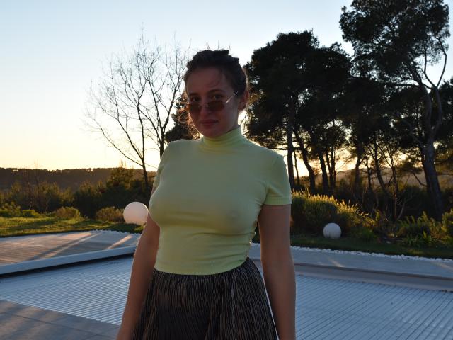 ZoeSchrute stands outside in a yellow turtleneck, showing her small tits, with a striped skirt, and round glasses, at sunset, with trees in the background.