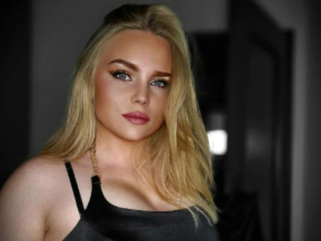 CherryDeluxe-hot has blonde hair, blue eyes, and is wearing a black tank top showing her tits. She's looking at the camera with pink lipstick and a serious expression.