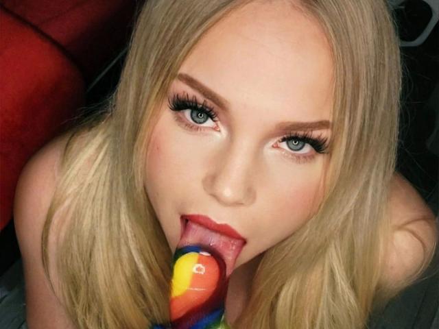 CherryDeluxe-hot with blonde hair, blue eyes, and thick eyelashes, sucking on a colorful, spiral-shaped dildo. Her tongue is out, lips parted, with a sexy, seductive look.