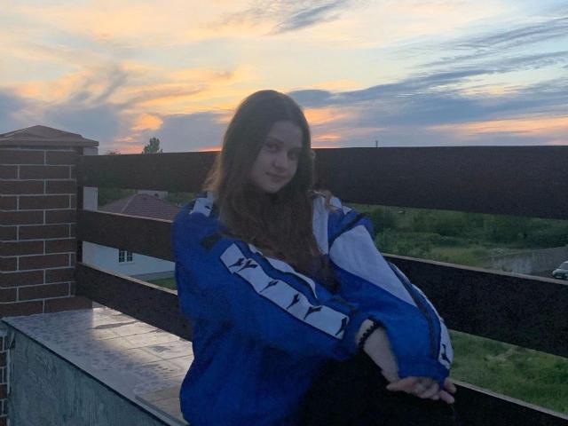 BellaMuse-hot with long brown hair, wearing a blue and white track jacket, sitting on a balcony at sunset, hands resting on her knees, smiling softly.