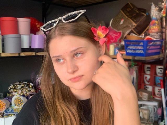 BellaMuse-hot with long brown hair, blue eyes, and a pink flower in her hair, wearing sparkly sunglasses, touching her ear, in a store with colorful containers.