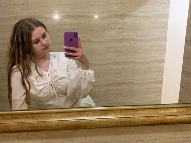 BellaMuse-hot with wavy brown hair, white blouse, and purple phone, taking a mirror selfie. Boobs slightly visible under blouse. Marble wall in background.
