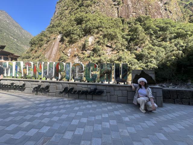 ElizaCanterville sits on a stone sidewalk with a mountain and forest behind her, wearing a white hat and patterned jacket, in front of colorful, graffiti-style letters.