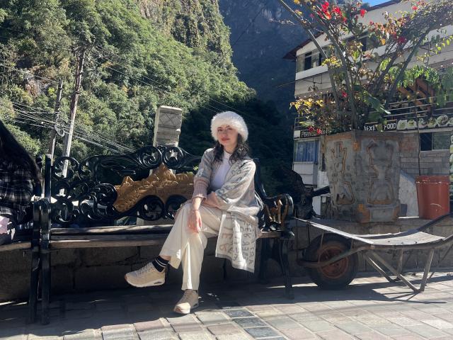 ElizaCanterville with fluffy white hair, wearing a silver patterned dress, white pants, and white sneakers, sitting on a black ornate bench, sunny outdoor setting, red and yellow leaves.