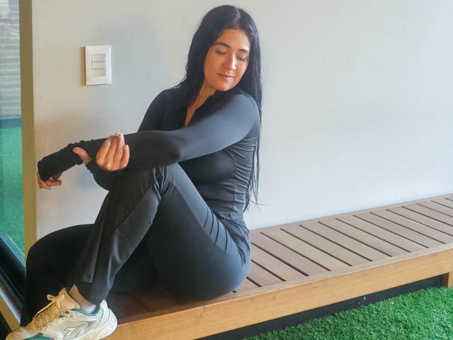 NicoColeman, with long black hair, sits on a wooden bench, stretching in a black athletic outfit, showing her ass, wearing white sneakers, eyes closed, in a relaxed pose.