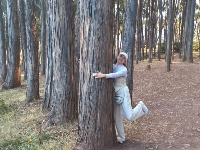ElizaCanterville is leaning against a tall, rough-boled tree, wearing a white headscarf and light gray clothes, with one leg kicked up in a forest.