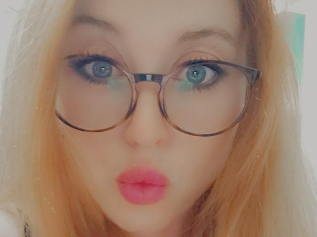 AliceNova, con lunghi capelli neri, occhiali, cuffie rosa con orecchie da gatto e rossetto rosa, sorride alla fotocamera, mostrando il seno, un tatuaggio sul braccio destro e un'illuminazione rosa..