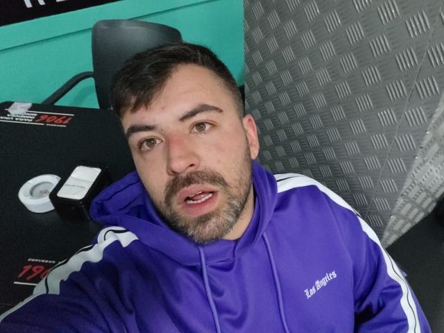 Vio69x, a bearded man with short brown hair, wearing a purple Adidas hoodie, sits with a slightly open mouth, looking at the camera. His face is slightly flushed.