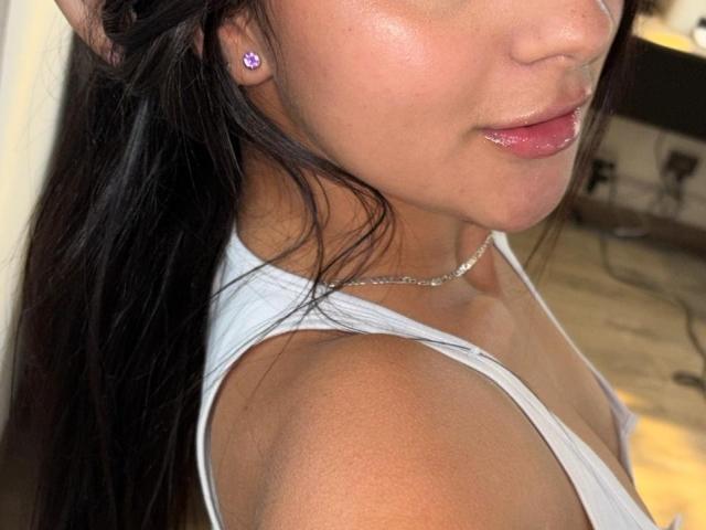 AlessiaFerry's face, showing shiny pink lips, a silver necklace, white tank top, and dark hair. She's got purple earrings and a subtle tan. Boobs not visible.