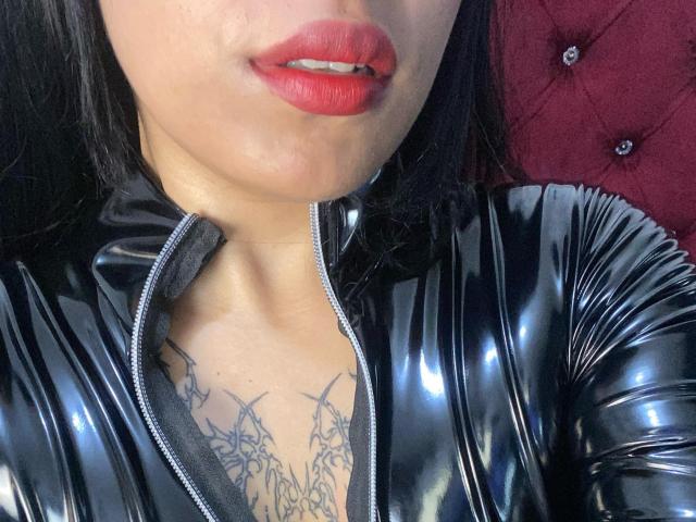 La tenue en latex noir brillante de ZaraBlaaze, avec son rouge à lèvres rouge et son tatouage visible sur la poitrine, constitué de fines lignes noires, sur un fond rouge texturé. Sa fermeture éclair est partiellement ouverte..