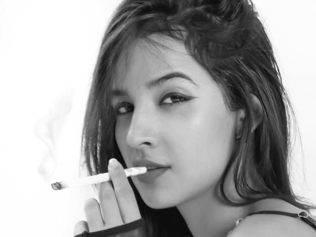 LeenaBrown with long dark hair, holding a cigarette in her right hand, smoke curling upwards, looking to the side with a slight smirk, black and white image.