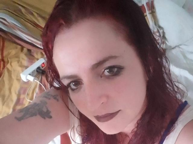 IsaWild-hot with red hair, tattoo on right arm, dark eyeliner, and dark lipstick, looking up with a neutral expression. Boobs partially visible, lying on a bed.