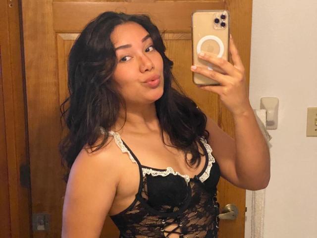 Ashlley69, avec ses cheveux noirs ondulés, ses lèvres pulpeuses, porte de la lingerie en dentelle noire avec des bords blancs, tient un téléphone, prend un selfie devant un miroir, montrant sa petite poitrine et son joli visage..