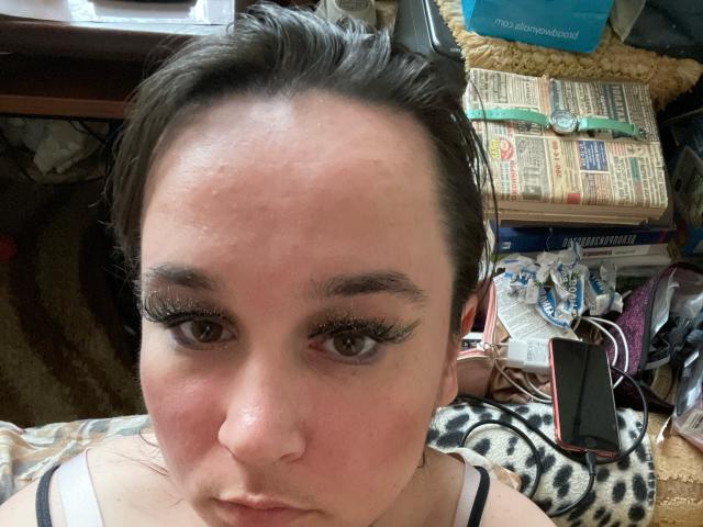 SaliBerry with dark hair, wearing black straps, has a pimple on her forehead, thick black eyelashes, and a neutral expression, lying on a leopard-print blanket.